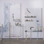 Photograph of artwork installations, two canvases hand on the wall, both are white with abstract markings on them and several small plastic structures stand on the floor in front of them, they are plastic poles standing at different heights