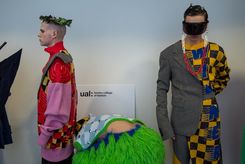 Male models waiting backstage, the model on the left wears a pink and red jacket and the model on the right wears a grey and yellow checked coat