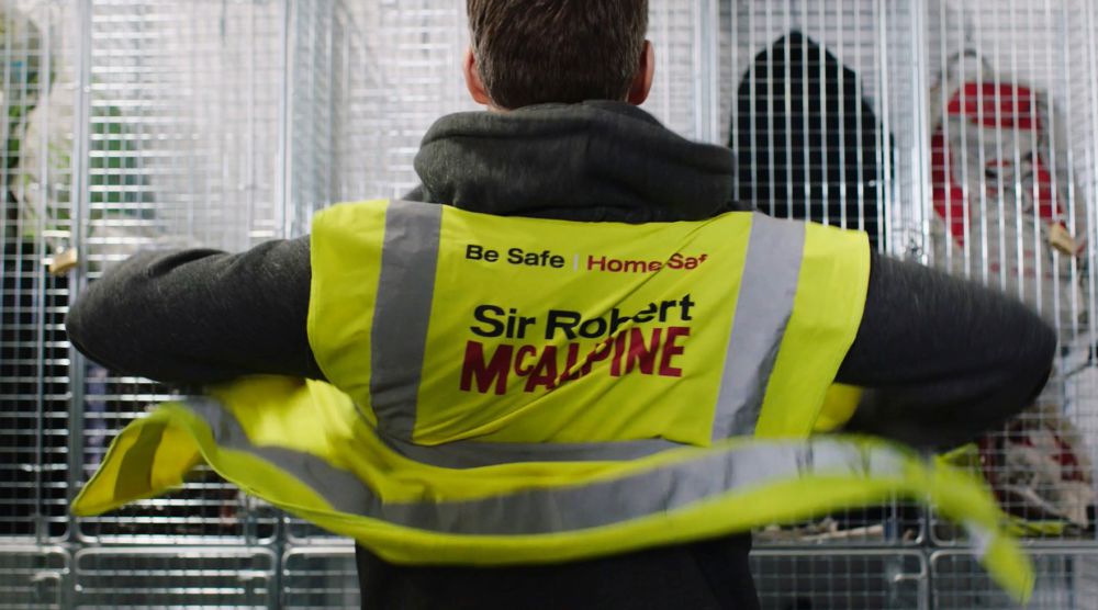 Still from a Health and Safety film showing a man facing a white locker, displayer the back of his Sir Robert McAlpine high-vis jacket. 