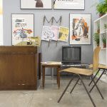 Photograph of an art installation that looks like a living area, there is a small dark wooden cabinet on the left with a box tv on a stand next to it on the right, with a wooden char in front and several framed prints hanging on the wall