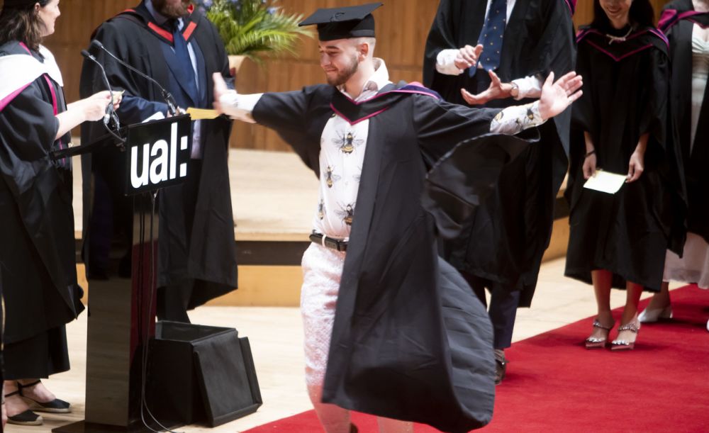 Graduate accepting his award and posing with his arms wide on stage