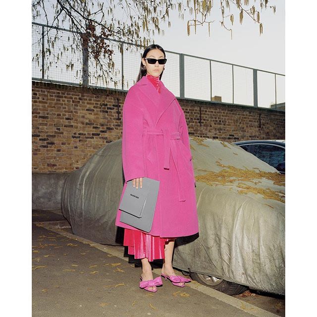 Photograph of a female model in a pink coat and sunglasses standing in a concrete setting