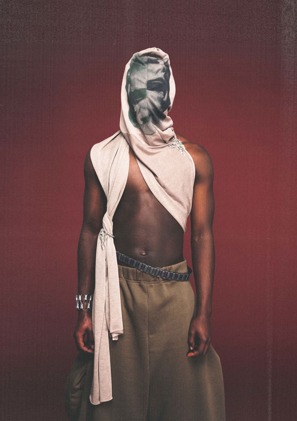 Male model with face covered by draping white scarf, in loose-fitting khaki trousers.