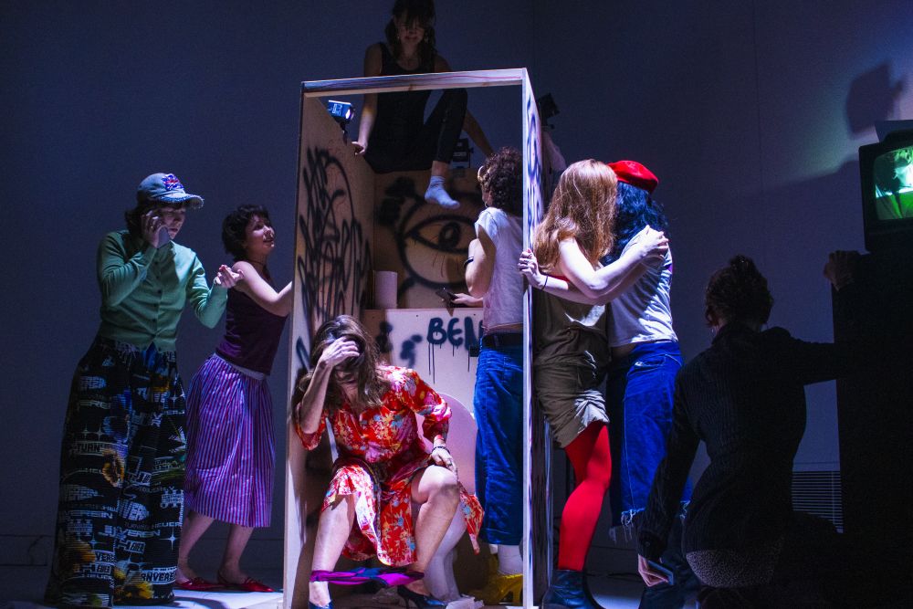 A group of performers on a dark blue stage wearing casual clothing, milling around and on top of a cubicle that has graffiti and a toilet, in which one performer is seated head in hands, knickers at ankles. Two performers are kissing to the right of the cubicle,
