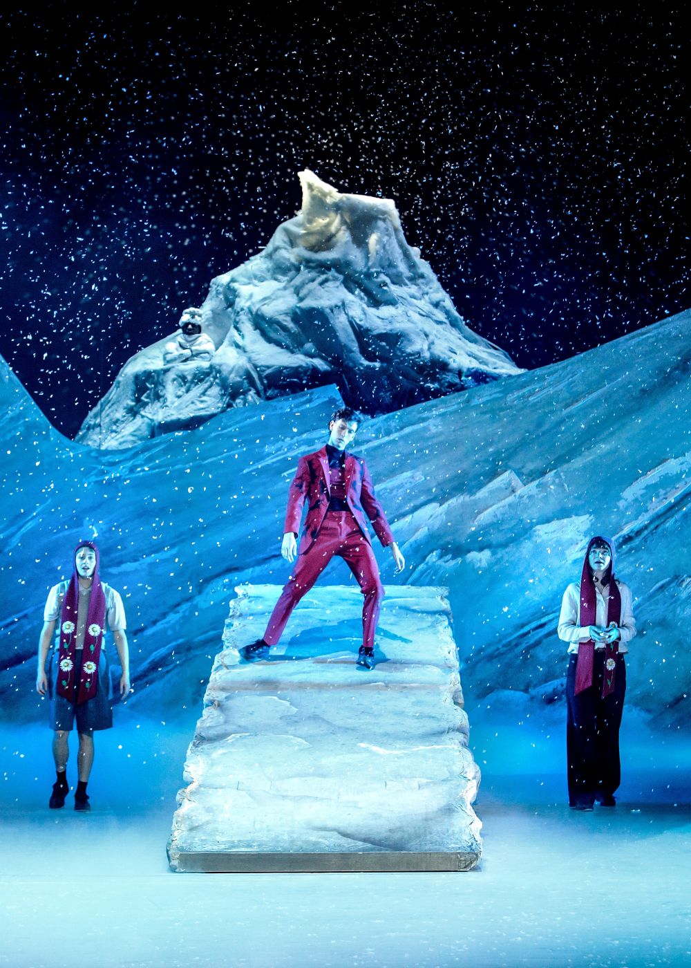 A theatrical scene set on a snowy, icy landscape with a large sculpted mountain backdrop. Artificial snow falls as three performers stand on stage. One performer in a bright red suit stands centered on an elevated icy platform, while two others stand on either side wearing winter-themed costumes and red sashes. The lighting creates a cold, wintry atmosphere.