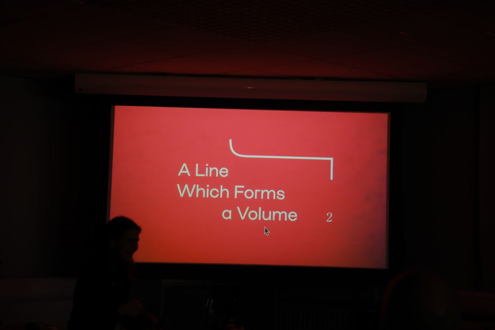 Red branding for A Line Which Forms A Volume projected onto the screen, with silhouettes.