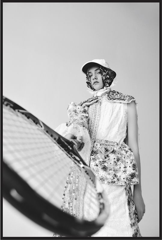 Black and white photograph of a model wearing a white hat and dress holding a tennis racket at the camera