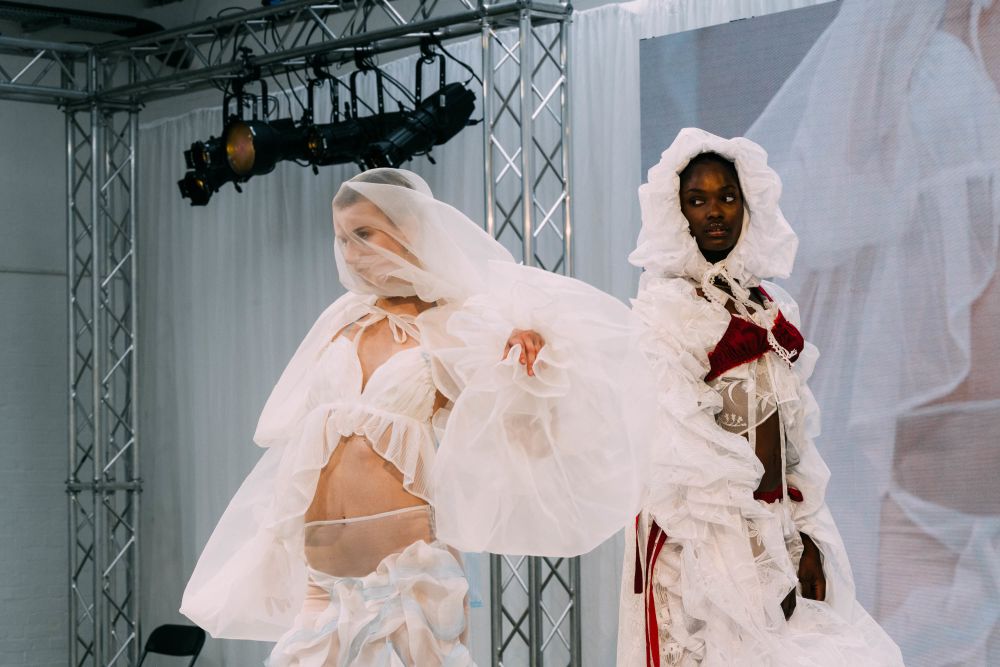 Two models on stage, wearing white outfits, with the model on the right also wearing a red bralette on top