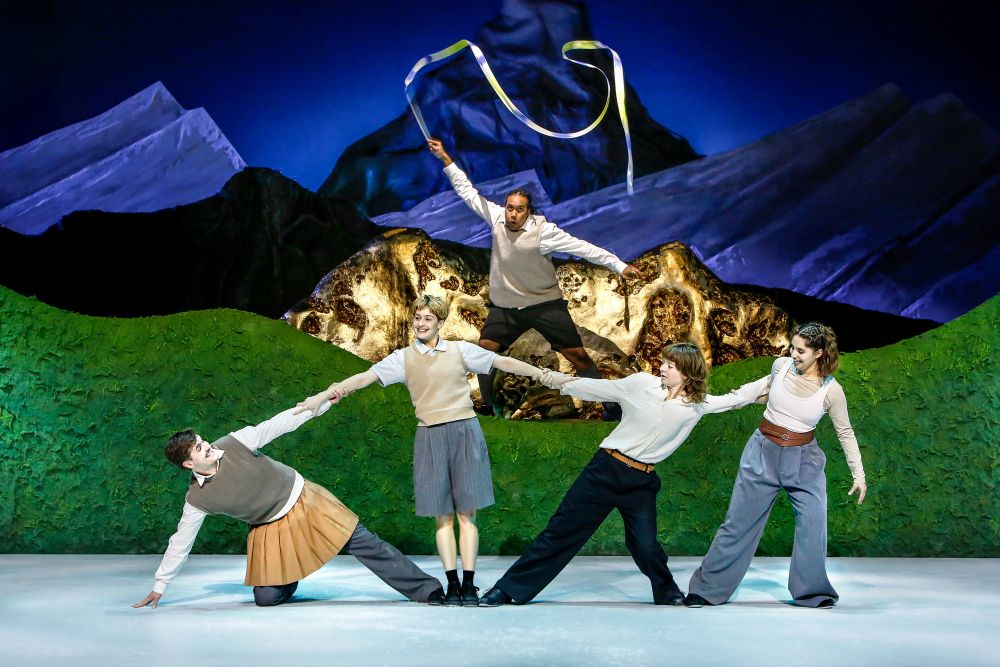 Five performers pose dynamically onstage in front of a painted mountain backdrop and green hillside set. One performer stands elevated at the back, holding a long ribbon overhead. The others create a chain-like formation, leaning and stretching while dressed in school-uniform‑style outfits. The set and costumes create a whimsical, storybook feel.