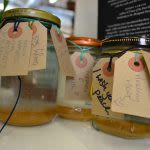 Photograph of a collection of jars with a small amount of honey-like liquid in the bottom, each one has a brown paper tag tied around the top 