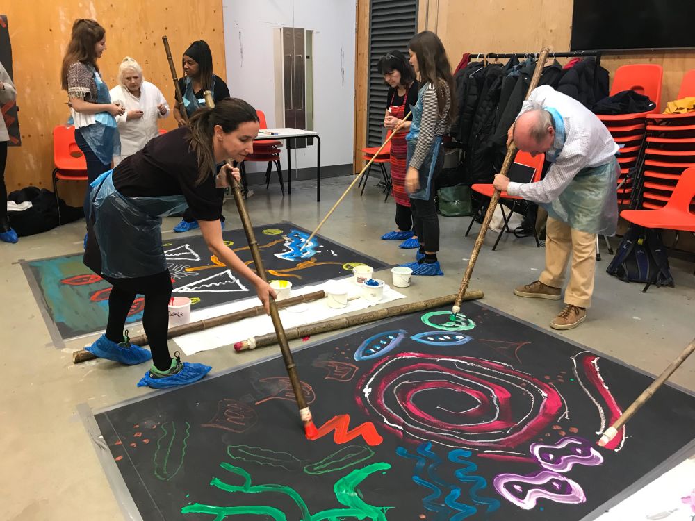 People painting with large sticks onto black canvas on floor