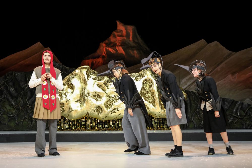 Four performers stand on an indoor stage against a dramatic mountainous backdrop with golden, textured scenery in the center. One performer stands to the left wearing a red scarf with decorative floral symbols, while three others on the right bend forward slightly, dressed in dark outfits with sculptural headpieces suggestive of long beaks or stylised animals.