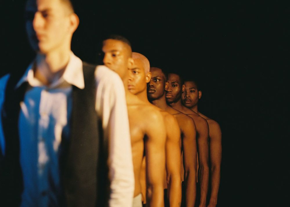 White male model in suit standing in front of a line of topless black men
