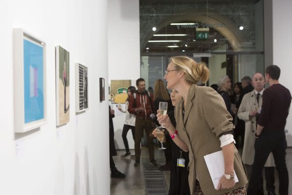 Photograph of visitors looking at art hung on the wall of a gallery, one woman is leaning forward to take a closer look at an artwork