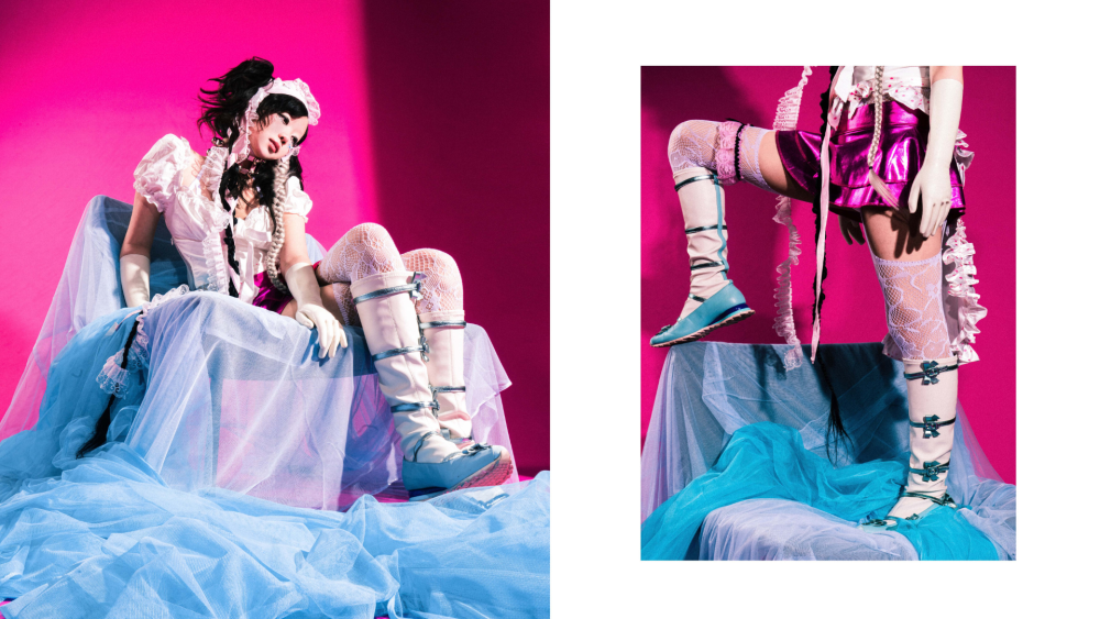 A model dressed in a pink shiny mini skirt, white cropped blouse and frilly bonnet sits on a chair draped in blue tulle. The studio backdrop is hot pink. 