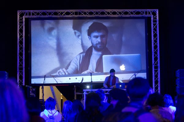 A photograph from behind a crowd watches someone DJ on stage, an image of him projected onto the screen behind him 