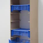 Photograph of an art installation detail, a wooden drawer frame hung on the wall, suspended slightly off the ground, with three electric blue plastic fruit crates slotted into it 
