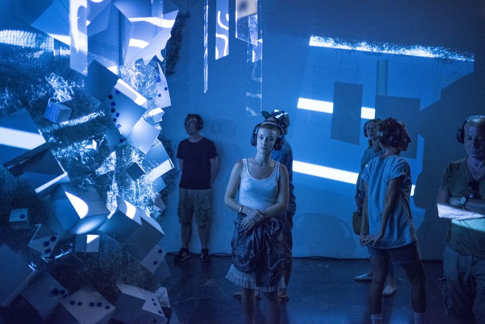 A woman is stanading in front of an installation, she is wearing headphones