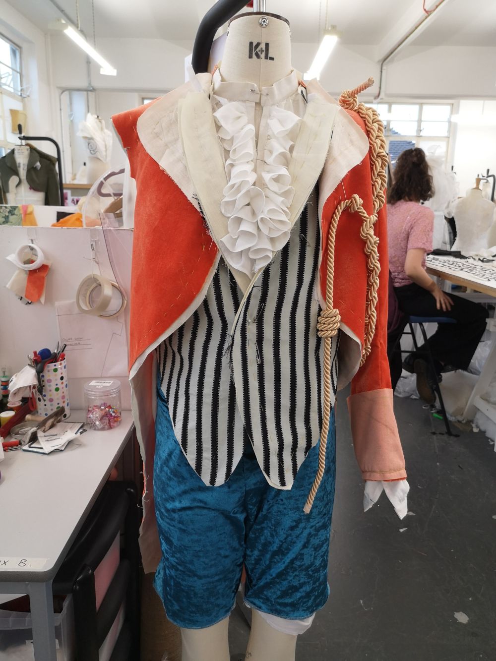 Photo of a partially made costume on a mannequin. The costume is made up of a red velvet jacket, striped waistcoat and blue trousers which are cropped at the knee. 