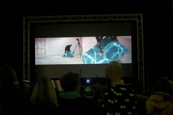 A photograph from behind a crowd watching a large screen above a stage as someone performs with a flat, square blue object on a split screen projection