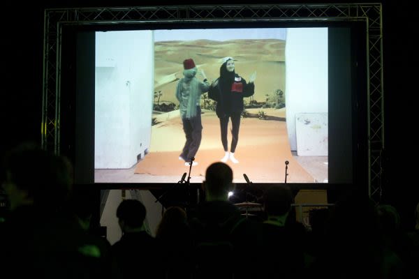 A photograph from behind a crowd watching a large screen showing two women dancing 
