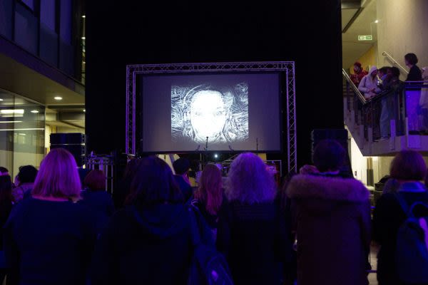 A photograph from behind a crowd watching a large screen above a stage as someone performs