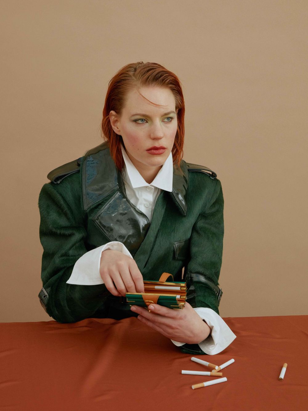 Ginger lady in green trench coat sitting at a table with a box of cigarettes