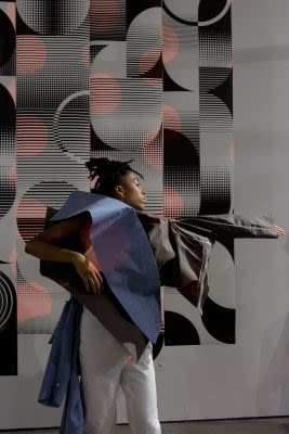 Photograph of someone performing, wearing a garmet with a large protrusion over the shoulder, stood infront of an abstract geometric pattern on the wall