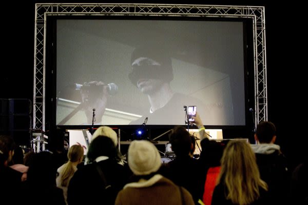 A photograph of a crowd from behind, watching someone perform on stage, a large screen behind showing a man with his mouth and nose covered by a mask