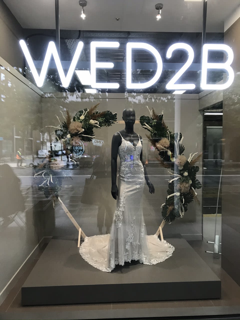 A photo of a mannequin wearing a long white dress in the window of a shop called WED2B.
