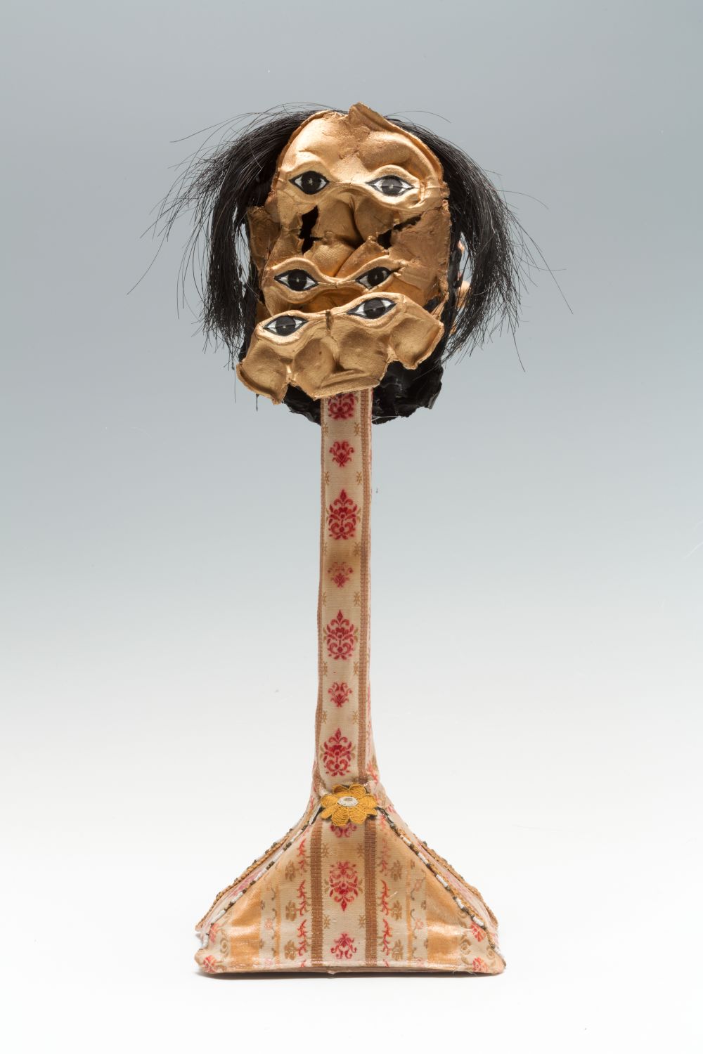 A mixed media sculpture of a figure with multiple sets of eyes and hair. There is a patterned motif on the upright body of the sculpture 
