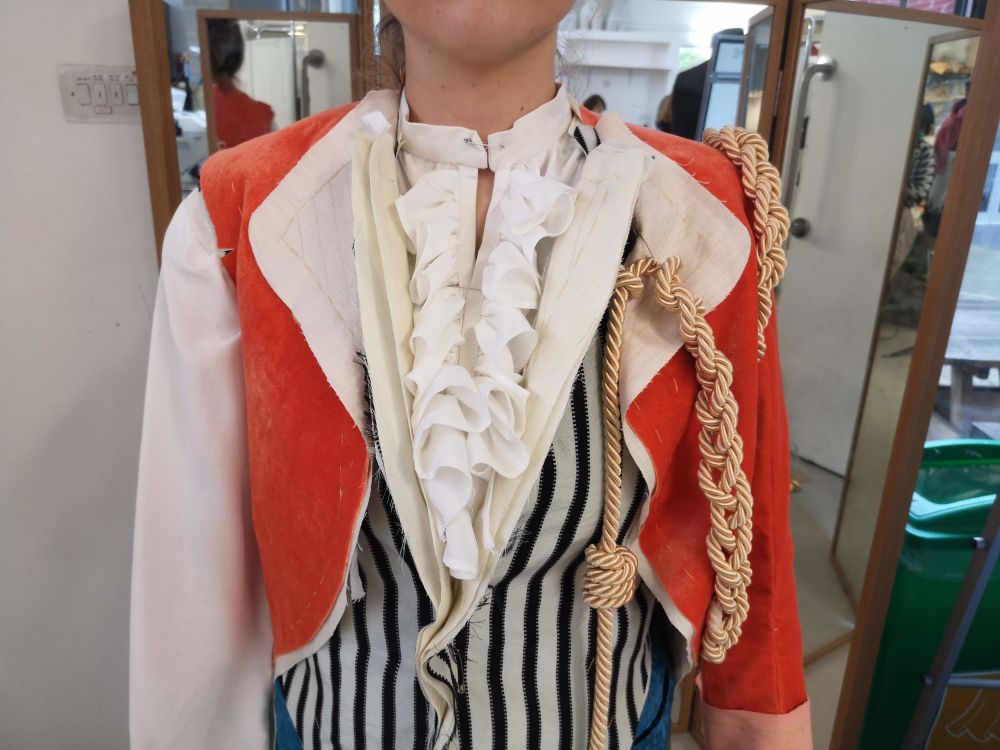 Photo of a partially made red velvet jacket, worn over a horizontal-striped waistcoat, with gold rope details. 