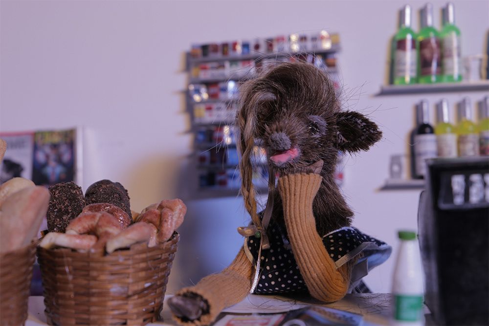 A female model of a deer, taken as a still from a stop motion animated film, sits in a bakery alongside some pastries.