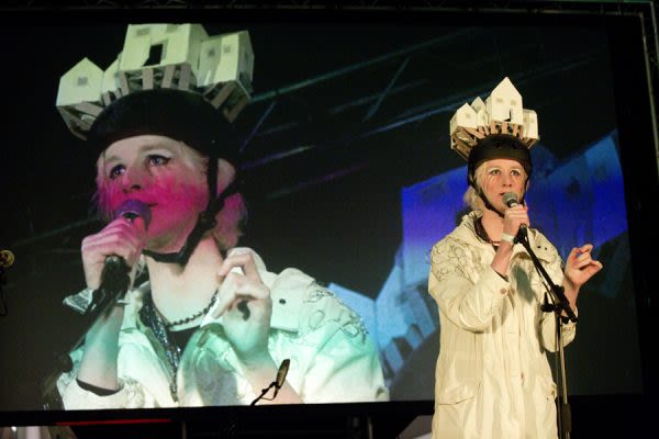 Photograph of someone performing on stage, wearing a hat made of miniature houses