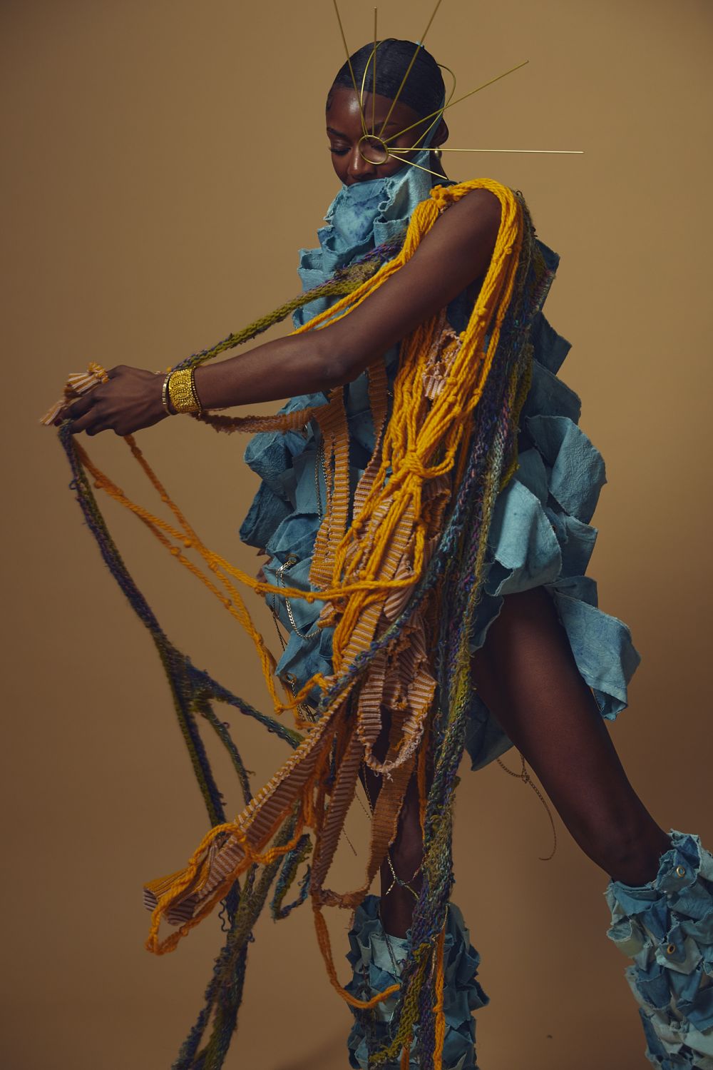 Model wearing a sculptural blue outfit with ruffled textures and long woven orange and green yarn strands, accessorized with a gold headpiece radiating outward from the face, set against a beige background.