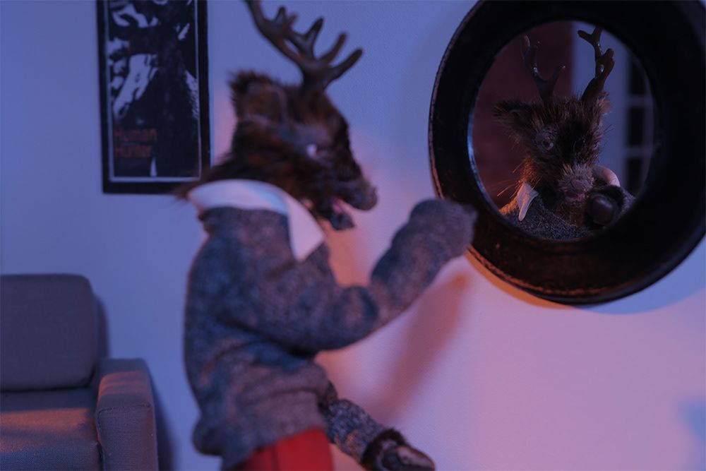 A male model of a deer, taken as a still from a stop motion animated film, waves to another deer in a shop scene, looks in a mirror with a scary face looking back.