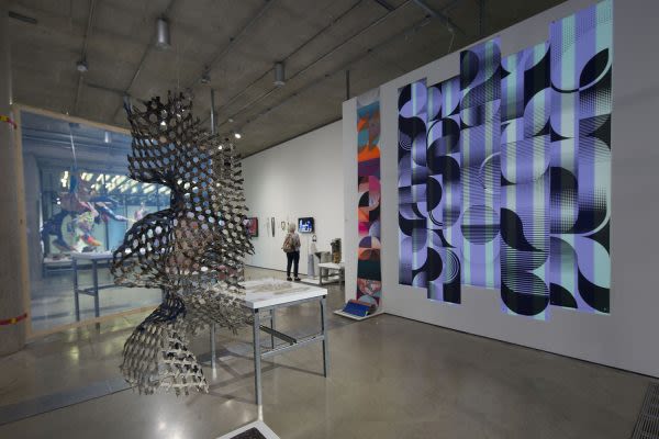  Photograph of the gallery with several pieces of artwork installed. a sculpture hangs from the ceiling in the middle and an image of a lilac and green absract pattern hangs on the wall behind it
