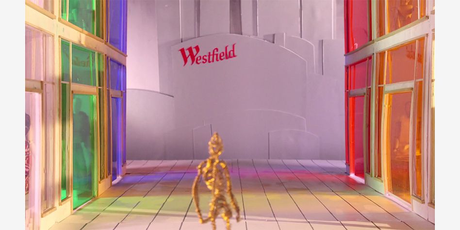 film still of human figure made out of metal wire whilst standing in multicoloured shopping centre looking at sign reading Westfield