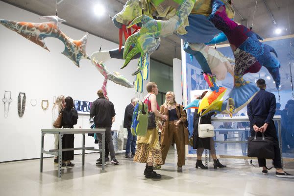 Photograph of people in the gallery socialising and looking at the artwork, a soft sculpture hangs from the ceiling in the foreground and the photograph is taken from a low angle 