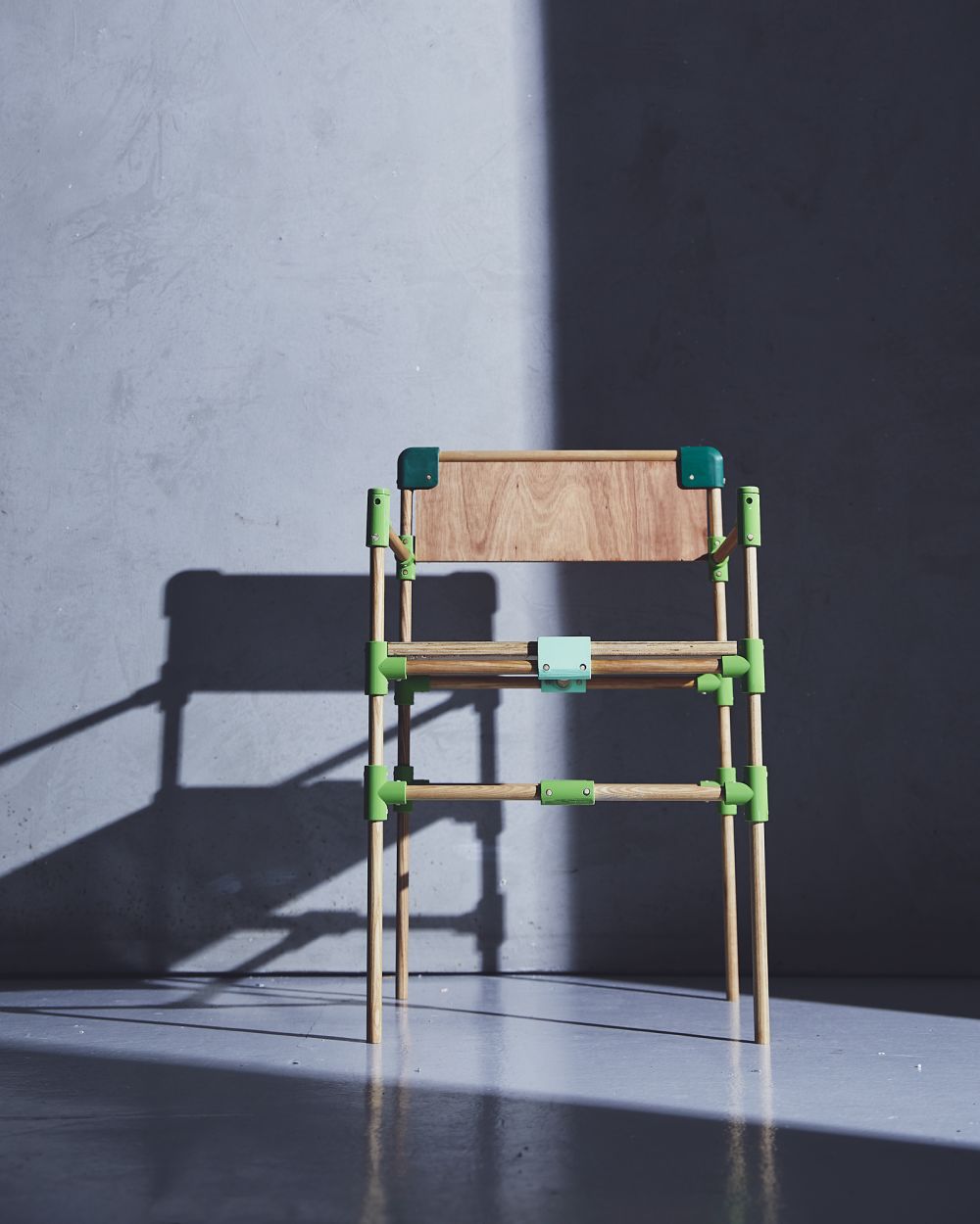 A wooden chair connected by green plastic joints shot against a concrete wall with harsh light from the sun cascading across it
