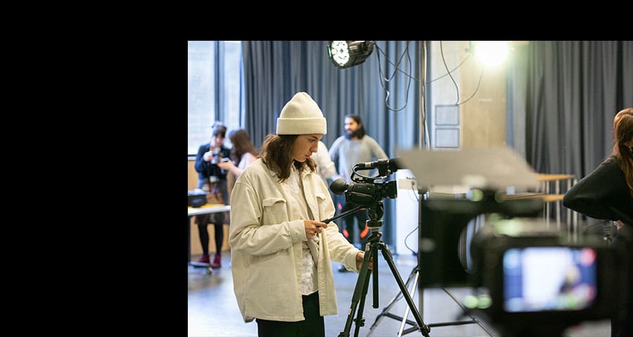 A white student in a white hat and jacket operates a camera as part of a class on camera skills