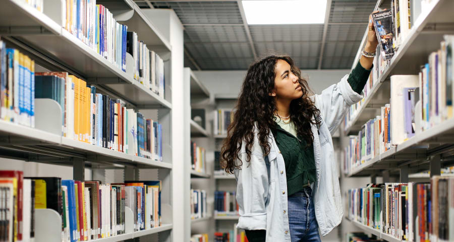 A woman stands in a library, examining books on the shelves.