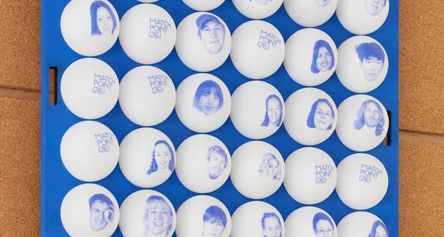 White tennis balls with blue faces and text saying 'Match Point 26' on them stuck to a blue board